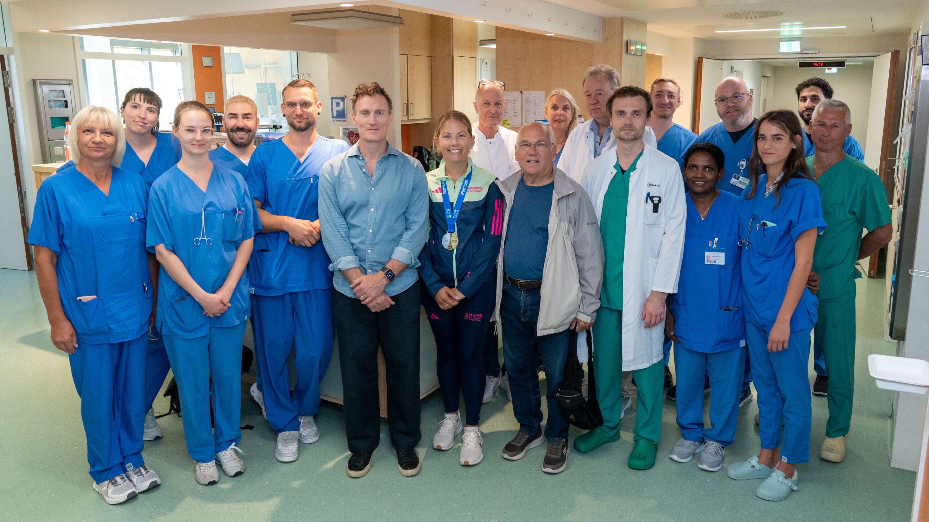 Eine Gruppe von medizinischem Personal in blauer Arbeitskleidung steht in einem Krankenhausflur. Sie lächeln und posieren gemeinsam für ein Foto. Im Vordergrund sind auch einige Besucher, die eng mit dem medizinischen Team verbunden sind.