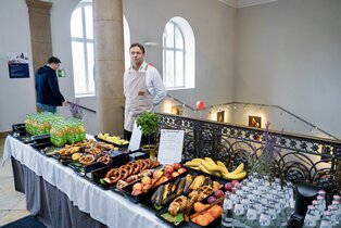Ein Buffet ist aufgebaut, das verschiedene Lebensmittel wie Brezeln, Obst und Getränke umfasst. Im Hintergrund steht ein Mann in weißer Kleidung, der das Buffet betreut, während eine andere Person auf der Treppe vorbeigeht.