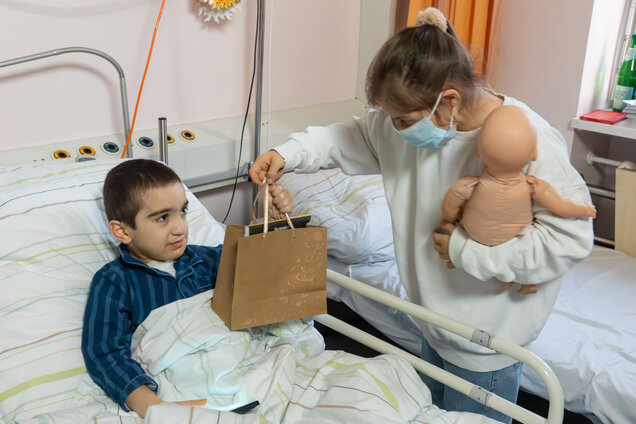 Ein Junge sitzt im Krankenhausbett, zugedeckt mit einer Decke. Ein Mädchen reicht ihm eine Geschenktüte. Sie hält eine Puppe in der anderen Hand und trägt eine Maske. Beide schauen sich an, wobei der Junge neugierig aussieht.