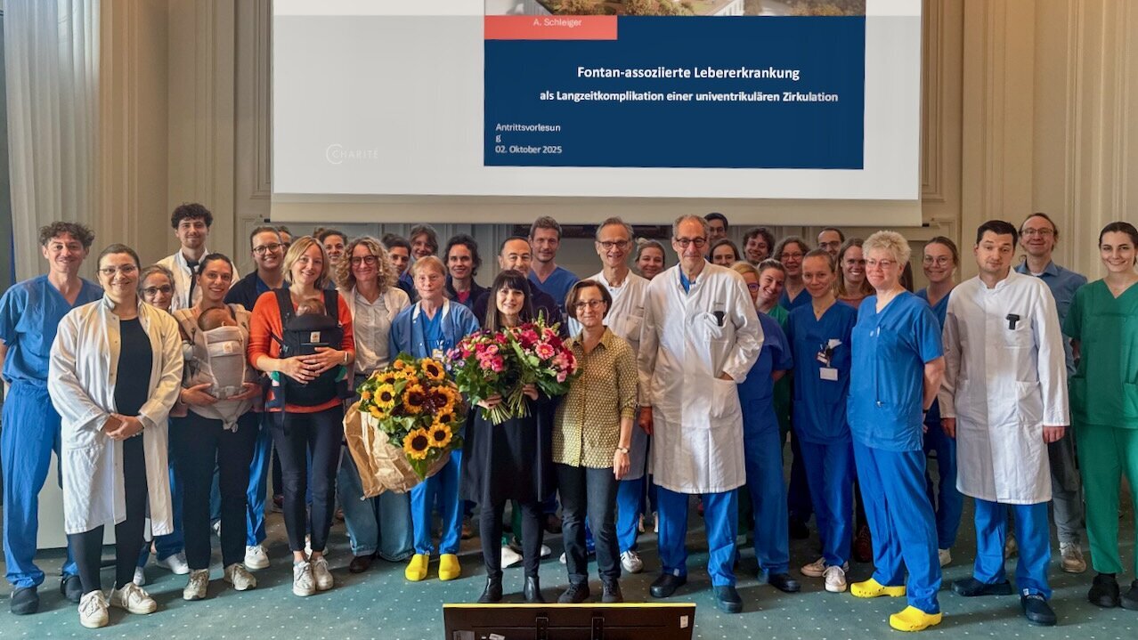 Eine Gruppe von medizinischem Personal steht lächelnd zusammen. Im Vordergrund hält eine Frau mit Blumenstrauß eine Präsentation über "Fontan-assozierte Lebererkrankung" in einem Raum des Deutschen Zentrums der Charité. Der Anlass ist eine Antrittsvorlesung.