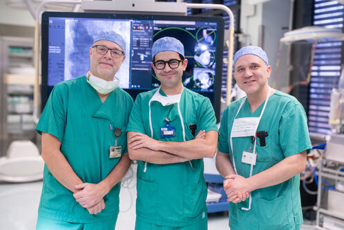 Drei Chirurgen in grünen OP-Kitteln stehen in einem modernen Operationssaal. Sie lächeln und haben chirurgische Hauben sowie Masken getragen. Im Hintergrund ist ein Bildschirm mit medizinischen Bildern zu sehen.