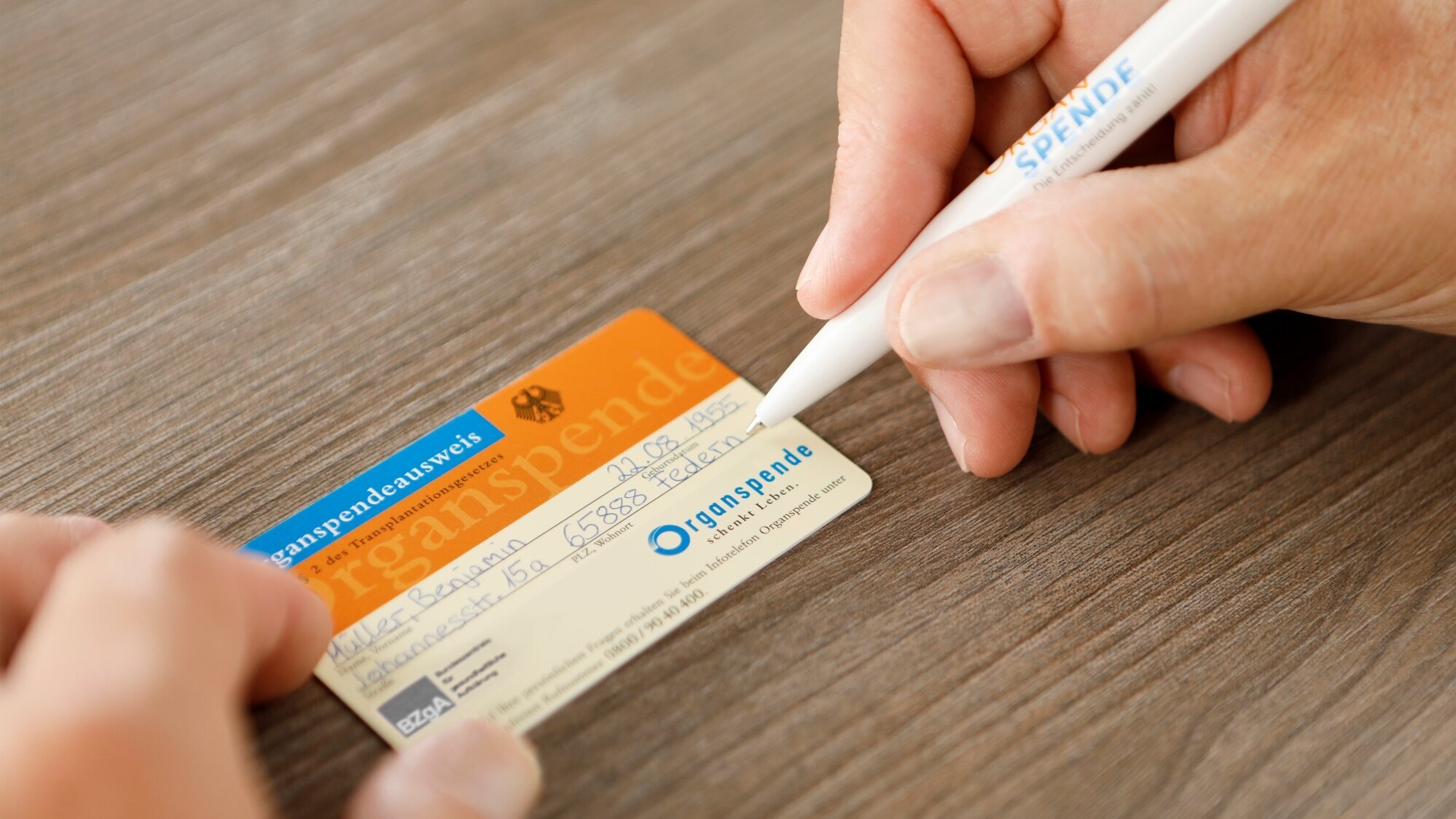 Eine Hand hält eine Spendenkarte, während eine andere Hand mit einem Stift darauf schreibt. Die Karte hat eine orangefarbene und blaue Farbgestaltung mit dem Wort "SPENDE". Der Hintergrund ist ein heller Holztisch.