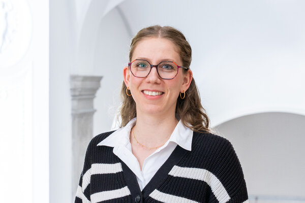 Ein Lächeln einer Frau mit langen, lockigen Haaren und einer Brille. Sie trägt einen schwarz-weißen gestreiften Pullover und ein weißes Hemd. Der Hintergrund ist hell und neutral.