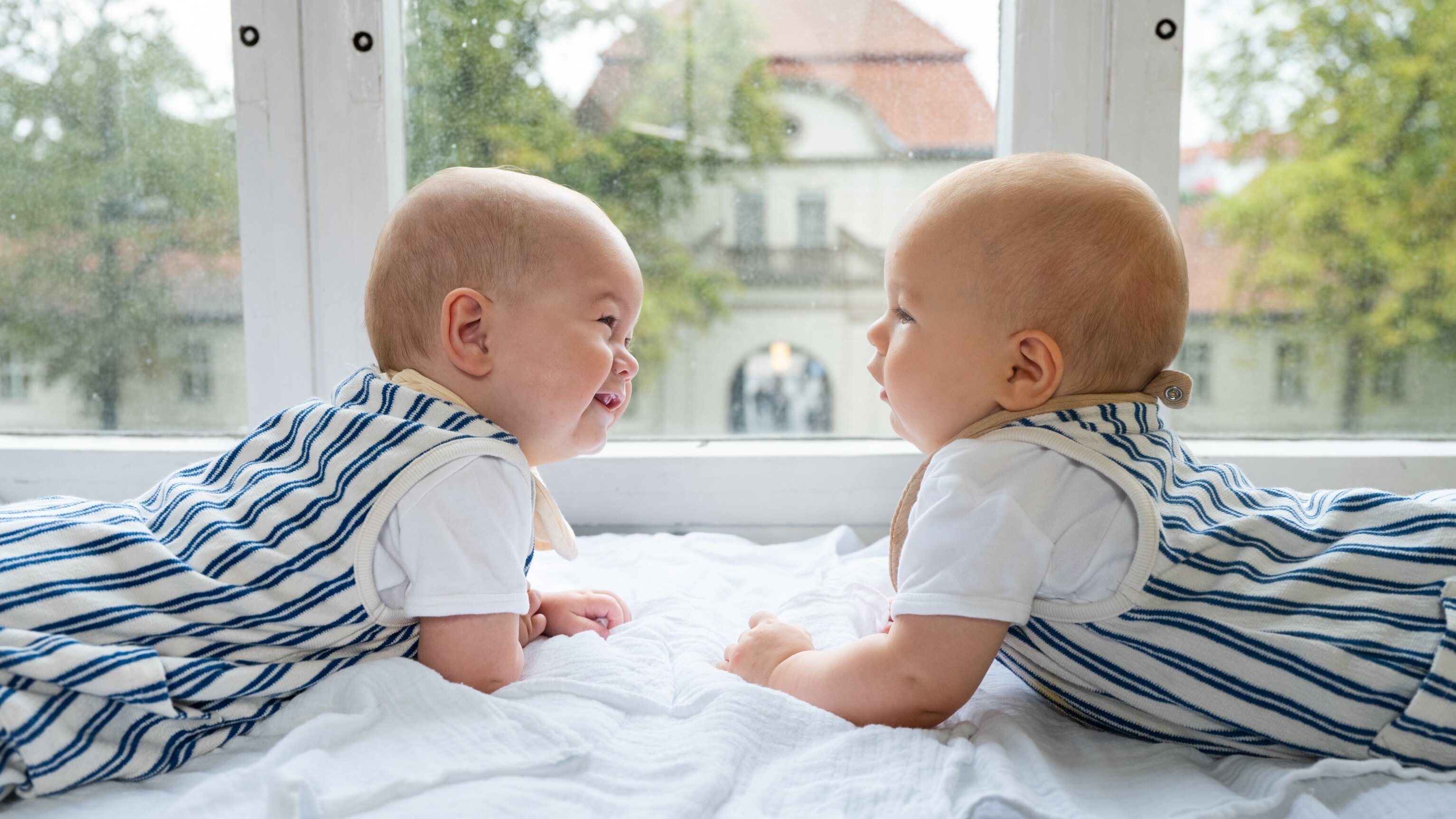 Zwei Babys liegen auf einer weißen Decke, lauschen und lächeln sich gegenseitig an. Sie tragen blau-weiß gestreifte Overalls. Im Hintergrund sind unscharfe Bäume und ein Gebäude sichtbar, während es draußen regnet. Sie sind in einem hellen Raum, der durch ein Fenster erleuchtet wird.