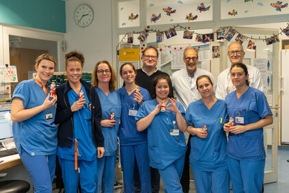 Eine Gruppe von medizinischem Personal, bestehend aus acht Frauen und mehreren Männern, steht in blauen Arbeitskleidung in einem Krankenhauszimmer. Alle halten kleine Figuren in der Hand. Im Hintergrund sind Krankenhausgeräte und ein Kalender sichtbar, was auf eine freundliche und kollegiale Atmosphäre hinweist.