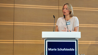 Eine blonde Frau steht am Rednerpult und spricht. Darunter ist ein Bildschirm mit ihrem Namen, „Marie Schafstedt“, und dem Datum „Berlin September 27, 2025“ zu sehen. Der Hintergrund besteht aus einer strukturierten Holzwand.