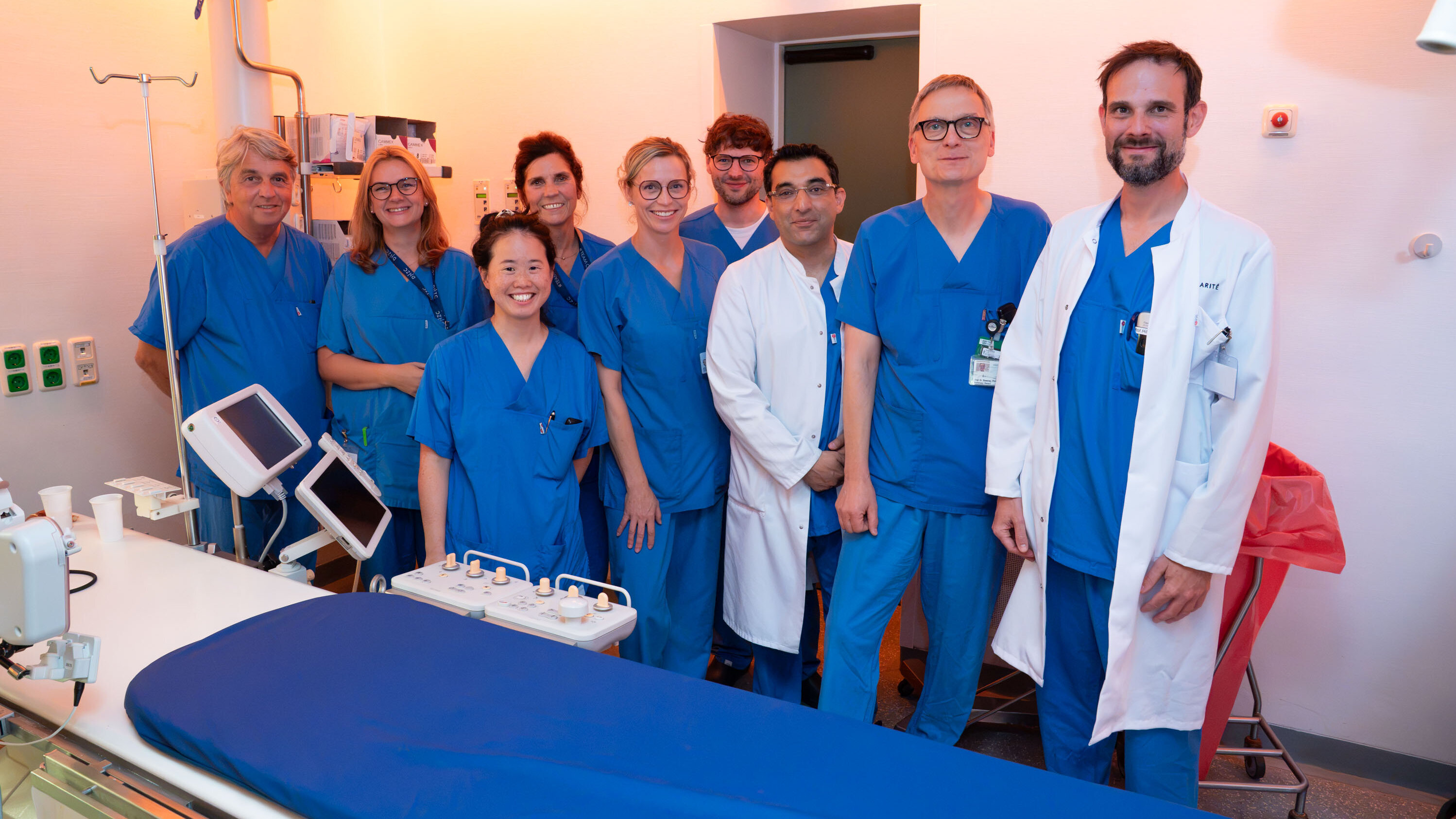 Eine Gruppe von zehn medizinischen Fachleuten in blauen Scrubs steht in einem Krankenhausraum. Sie lächeln und posieren für das Foto. Im Hintergrund ist ein Bett mit blauen Laken und medizinische Geräte sichtbar. Es herrscht eine freundliche Atmosphäre.