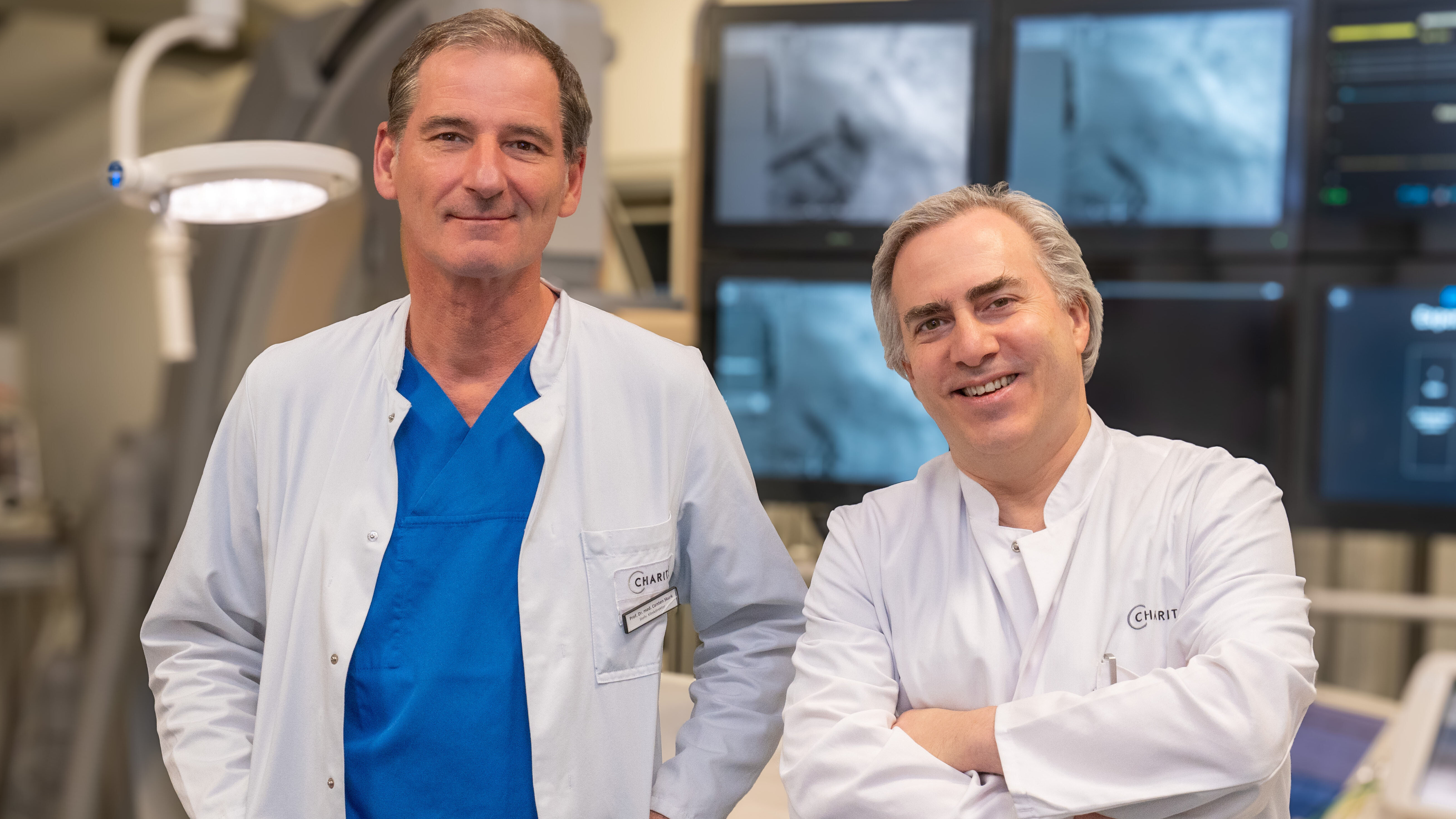 Zwei Ärzte in einem Operationssaal stehen nebeneinander und lächeln. Der erste trägt einen weißen Kittel und hat ein blaues Oberteil an, der zweite hat ebenfalls einen weißen Kittel. Im Hintergrund sind Monitorbildschirme zu sehen.