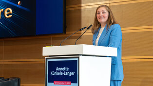 Eine lächelnde Frau in einem blauen Anzug steht an einem Rednerpult. Im Hintergrund ist eine Präsentation sichtbar, die verschiedene Himmelskörper zeigt. Der Name „Annette Künkele-Langer“ und das Datum „27. September 2025, Berlin“ sind auf dem Pult zu sehen.