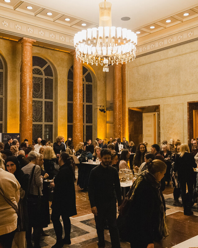 Eine belebte Veranstaltung in einem festlich beleuchteten Raum mit hohen Fenstern und marmorierten Säulen. Menschen stehen in Gruppen zusammen, unterhalten sich und genießen Getränke und Snacks. Ein großer Kronleuchter hängt von der Decke und trägt zur eleganten Atmosphäre bei.