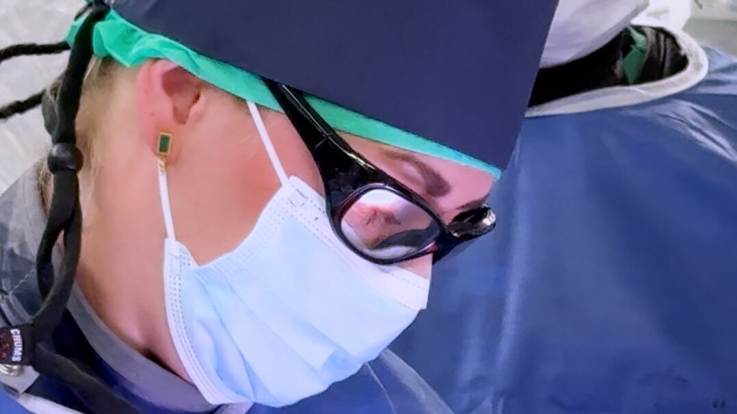 Zwei medizinische Fachkräfte in OP-Kleidung und Schutzmasken konzentrieren sich auf einen chirurgischen Eingriff. Die Person im Vordergrund trägt eine spezielle Brille und einen Operationshaube, während die zweite Person im Hintergrund ebenfalls Schutzkleidung trägt.
