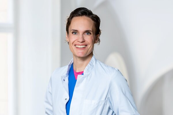 Eine freundlich lächelnde Person trägt einen weißen Laborkittel über einem blauen Oberteil. Sie steht in einem hellen Raum mit sanften, neutralen Farben. Die Person strahlt Professionalität und Zugänglichkeit aus.