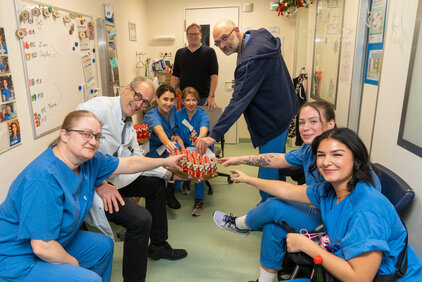 Eine Gruppe von sieben Personen in blauen Arbeitskleidungen sitzt und steht in einem Flur. Sie lächeln und zeigen auf einen Tisch mit kleinen Schokoladenfiguren. Die Atmosphäre ist freundlich und feierlich, was auf eine besondere Gelegenheit hinweist.