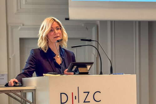 Eine blonde Frau mit Brille steht an einem Rednerpult und spricht in ein Mikrofon. Sie trägt einen dunkelblauen Anzug und schaut aufmerksam in die Richtung des Publikums. Das Pult trägt das Logo „D·I·Z·C“. Im Hintergrund ist eine Präsentation sichtbar.