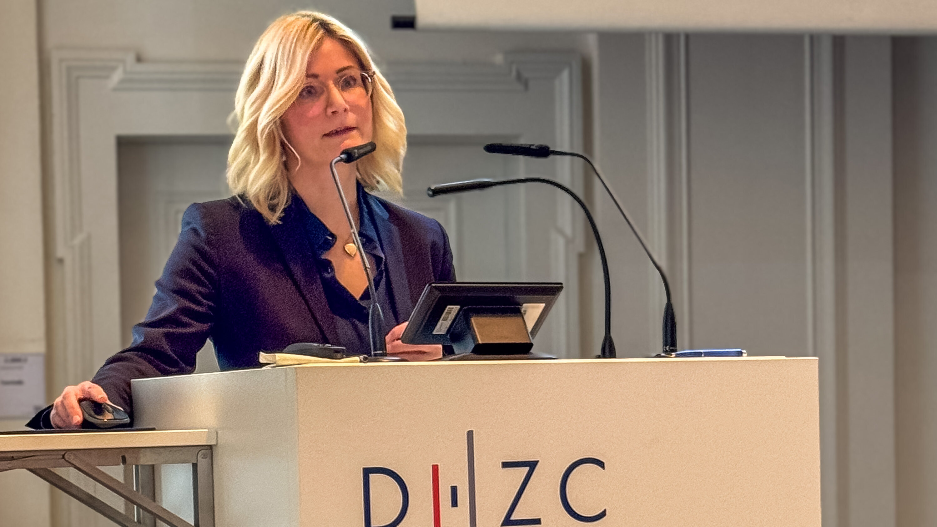 Eine blonde Frau mit Brille steht an einem Rednerpult und spricht in ein Mikrofon. Sie trägt einen dunkelblauen Anzug und schaut aufmerksam in die Richtung des Publikums. Das Pult trägt das Logo „D·I·Z·C“. Im Hintergrund ist eine Präsentation sichtbar.