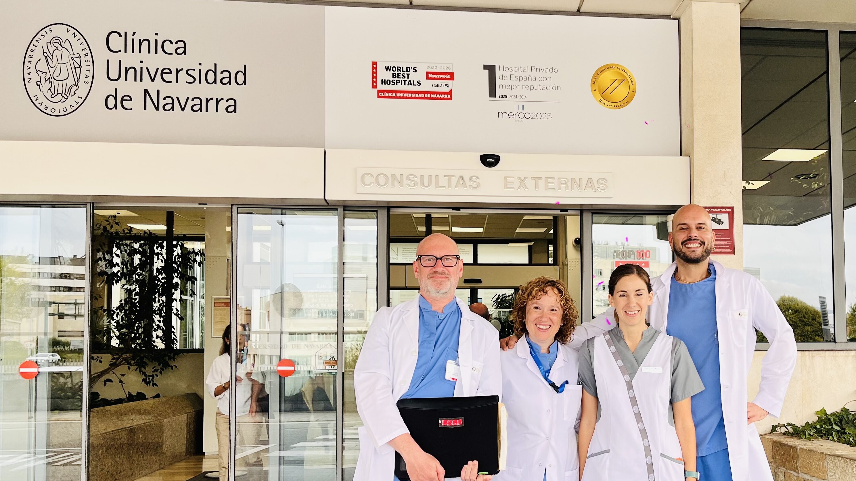 Das Bild zeigt vier medizinische Fachkräfte vor dem Eingang der Clínica Universidad de Navarra. Sie tragen weiße Kittel und lächeln freundlich in die Kamera. Im Hintergrund ist ein Schild mit dem Kliniknamen und dem Hinweis auf externe Konsultationen sichtbar.