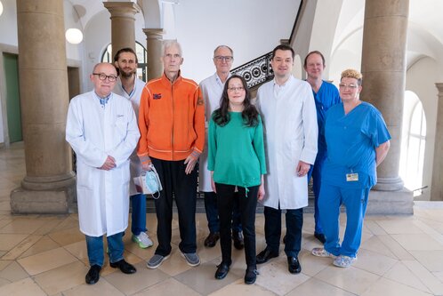 Eine Gruppe von neun Personen steht in einem Innenraum. Sie tragen verschiedene Kleidung, darunter medizinische Laborkittel und Berufsbekleidung. Im Vordergrund steht eine Frau in grüner Bluse zwischen Männern in Kitteln und einem orangefarbenen Oberteil. Der Raum hat hohe Fenster und eine elegante Treppe.