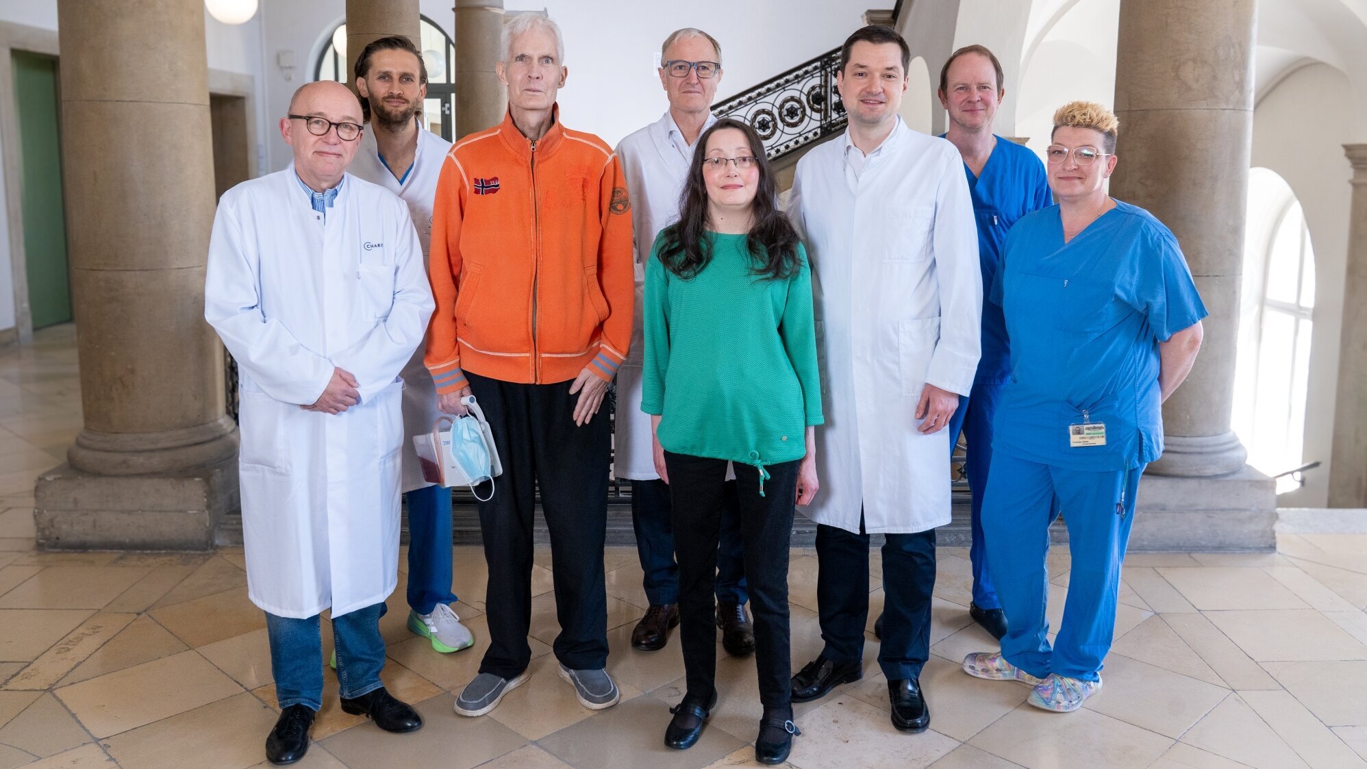 Eine Gruppe von neun Personen steht in einem Innenraum. Sie tragen verschiedene Kleidung, darunter medizinische Laborkittel und Berufsbekleidung. Im Vordergrund steht eine Frau in grüner Bluse zwischen Männern in Kitteln und einem orangefarbenen Oberteil. Der Raum hat hohe Fenster und eine elegante Treppe.