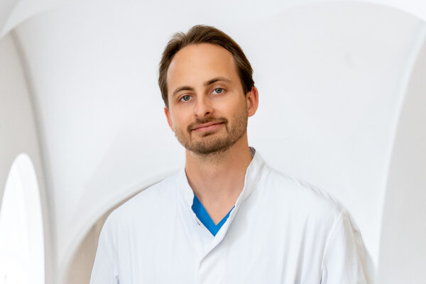 Ein Arzt in einem weißen Kittel steht vor einer hellen Wand. Er hat kurzes braunes Haar und trägt ein blaues Oberteil unter dem Kittel. Sein Gesicht zeigt ein freundliches Lächeln. Die Umgebung ist minimalistisch und hell gestaltet.