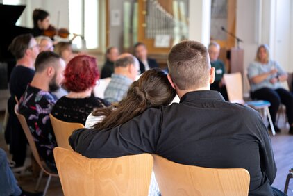 Eine Gruppe von Menschen sitzt in einem hellen Raum. Im Vordergrund sind zwei Personen zu sehen, die sich anlehnen. Im Hintergrund sind weitere Teilnehmer bei einer Diskussion oder Präsentation zu beobachten, während eine Person mit einem Instrument spielt.