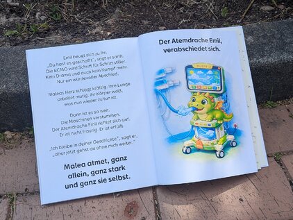 Das Bild zeigt ein geöffnetes Kinderbuch mit bunten Illustrationen. Auf der linken Seite sind Textzeilen zu sehen, während die rechte Seite einen niedlichen Drachen namens Emil zeigt, der an ein medizinisches Gerät angeschlossen ist. Der Drache erscheint freundlich und einladend.