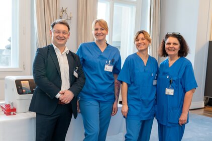 Eine Gruppe von vier Personen steht in einem hellen Raum. Drei Frauen tragen blaue Arbeitskleidung, während ein Mann in einem Anzug daneben steht. Alle lächeln und wirken freundlich. Im Hintergrund steht ein medizinisches Gerät auf einem Tisch.
