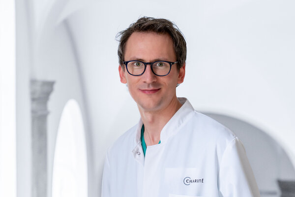 Ein Arzt in weißem Kittel steht lächelnd in einem hellen Raum. Er trägt eine Brille und zeigt eine freundliche Ausdrucksweise. Der Hintergrund ist unscharf, wodurch sein Gesicht und seine Kleidung hervorstechen.