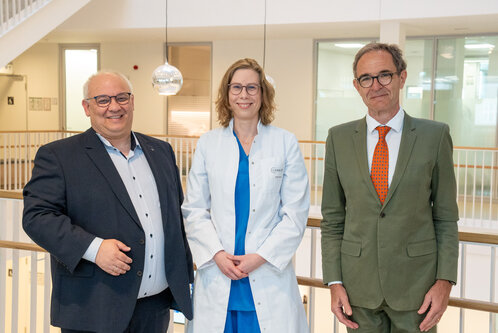 Drei Personen stehen in einem modernen Innenraum. Eine Frau in einem weißen Kittel vermittelt einen medizinischen Kontext, während die anderen beiden Personen in formeller Kleidung gekleidet sind. Alle lächeln und blicken in die Kamera.