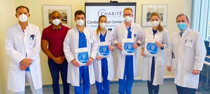 Eine Gruppe von sieben Personen in weißer Berufsbekleidung und Mundschutz steht zusammen. Sie halten Zertifikate in den Händen und lächeln. Im Hintergrund ist ein Bildschirm mit dem Logo der Charité und einer Präsentation zu sehen.