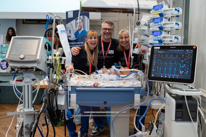 Drei Personen stehen lächelnd vor einem Krankenhausbett mit medizinischen Geräten. Im Hintergrund sind Pflegekräfte und ein plakatiertes medizinisches Thema zu sehen. Die Atmosphäre wirkt freundlich und unterstützend.