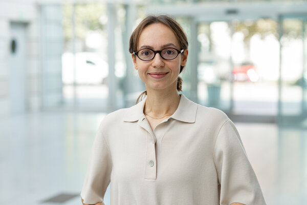 Eine junge Frau mit braunen, schulterlangen Haaren und einer hellen Bluse steht lächelnd in einem modernen Innenraum. Sie trägt eine Brille und blickt freundlich in die Kamera. Der Hintergrund zeigt unscharfe Fenster und helles Licht.
