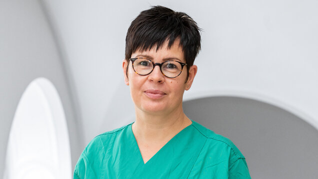 Frau in einem grünen Pflegehemd steht vor einem hellen, neutralen Hintergrund. Sie trägt eine Brille und hat kurzes, dunkles Haar. Ihr Gesichtsausdruck ist freundlich und professionell. Die Umgebung ist einfach und modern gestaltet.