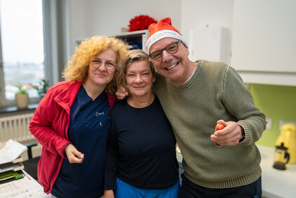 Drei Personen stehen eng beieinander und lächeln. Die Frau links trägt eine rote Jacke und hat lockige Haare, die Frau in der Mitte hat dunkles, kurzes Haar. Der Mann rechts trägt einen Weihnachtsmann-Hut und hält ein rotes Objekt in der Hand. Sie scheinen eine fröhliche Atmosphäre zu genießen.