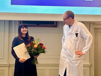 Eine lächelnde Frau hält einen Blumenstrauß und ein Zertifikat in der Hand. Neben ihr steht ein Mann in einem weißen Kittel, der freundlich schaut. Die Szene findet bei einer Veranstaltung zur Anerkennung oder Feierlichkeit statt.