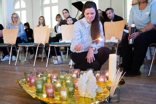 Eine Frau zündet mit einem langen Streichholz eine Kerze in einem Arrangement aus mehreren bunten Teelichtern und einer größeren weißen Kerze. Im Hintergrund sind mehrere Menschen in einer Sitzreihe zu sehen, die die Szene aufmerksam beobachten.