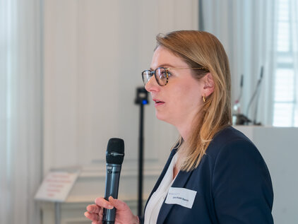 Eine Frau mit Brille hält ein Mikrofon und spricht. Sie trägt einen dunklen Blazer und hat schulterlange, blond gefärbte Haare. Im Hintergrund sind Fenster und ein Podium zu sehen, das ihre Präsentation unterstützt.