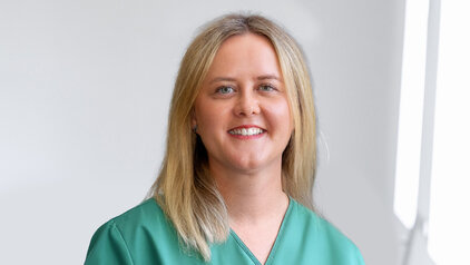 Ein lächelnde Frau mit blonden Haaren trägt einen grünen Kittel. Der Hintergrund ist hell und unscharf, sodass der Fokus auf ihrem Gesicht und ihrem freundlichen Ausdruck liegt. Die Aufnahme vermittelt eine positive und professionelle Atmosphäre.