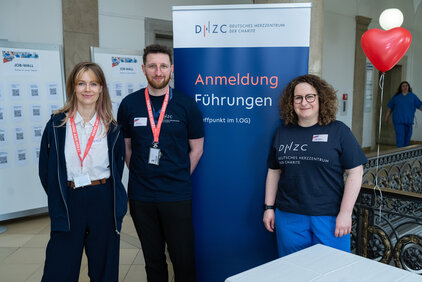 Drei Mitarbeitende stehen vor einem Banner des Deutschen Herzsuchzentrums. Der Text auf dem Banner informiert über Anmeldungen für Führungen. Im Hintergrund sind Plakate und eine Person in blauer Kleidung zu sehen.