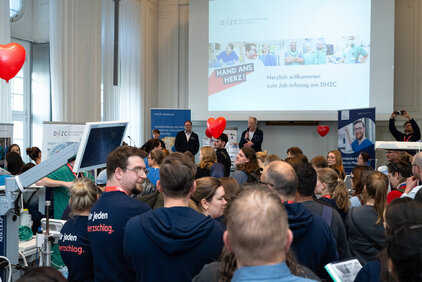 Eine größere Menschenmenge versammelt sich in einem Raum während einer Informationsveranstaltung zum Thema Jobangebote. Im Hintergrund sind Präsentationen und Informationsstände zu sehen, während Redner auf einer Bühne stehen. Rote Luftballons sind im Raum dekoriert.