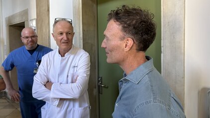 Drei Männer stehen in einem Flur. Einer trägt einen weißen Kittel, während ein anderer in blauer Arbeitskleidung gekleidet ist. Sie unterhalten sich freundlich. Im Hintergrund ist eine grüne Tür sichtbar. Der Raum hat helle Wände und große Fenster.