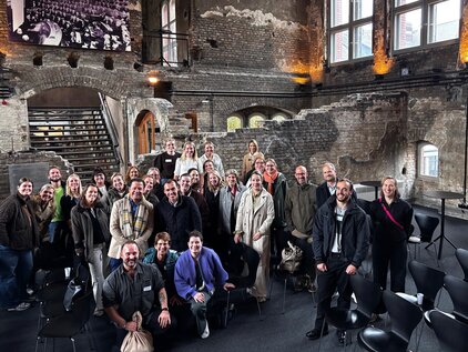 Eine Gruppe von etwa 35 Personen steht in einem großzügigen, lichtdurchfluteten Raum mit historischen Wänden. Die Teilnehmer lächeln und posieren für das Foto, einige tragen namensschilder. Im Hintergrund sind Fenster und Sitzgelegenheiten sichtbar.