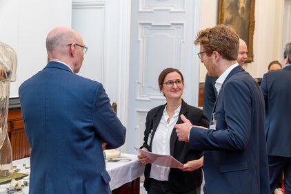 Drei Personen stehen in einer geschäftlichen Umgebung zusammen. Eine Frau hält Dokumente in der Hand und spricht mit einem Mann, während ein anderer Mann zuhört. Auf einem Tisch im Vordergrund sind verschiedene Objekte und Materialien zu sehen.