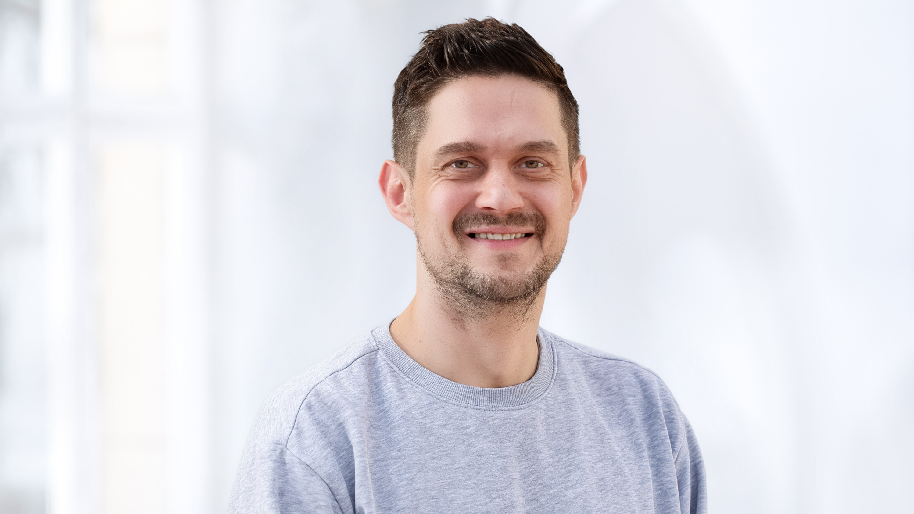 Ein lächelnder Mann steht vor einem hellen Hintergrund. Er trägt ein graues Sweatshirt und hat kurzes, dunkles Haar. Sein Gesicht zeigt Freude und Freundlichkeit.