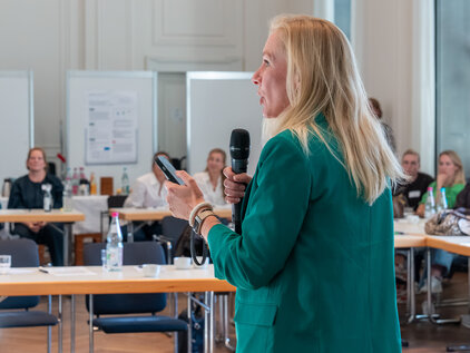 Eine Frau in einem grünen Blazer hält ein Mikrofon und spricht vor einer Gruppe von Zuhörern in einem Konferenzraum. Im Hintergrund sind mehrere Tische mit Getränken und Notizen zu sehen, während die Zuhörer aufmerksam zuhören.