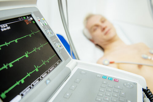 Ein Herzmonitor zeigt die Herzfrequenz und den Herzrhythmus eines Patienten an, der auf einem Bett liegt. Der Patient hat Elektroden an der Brust, während die Anzeige klare grüne Wellenlinien darstellt. Der Hintergrund ist unscharf und dezent gehalten.