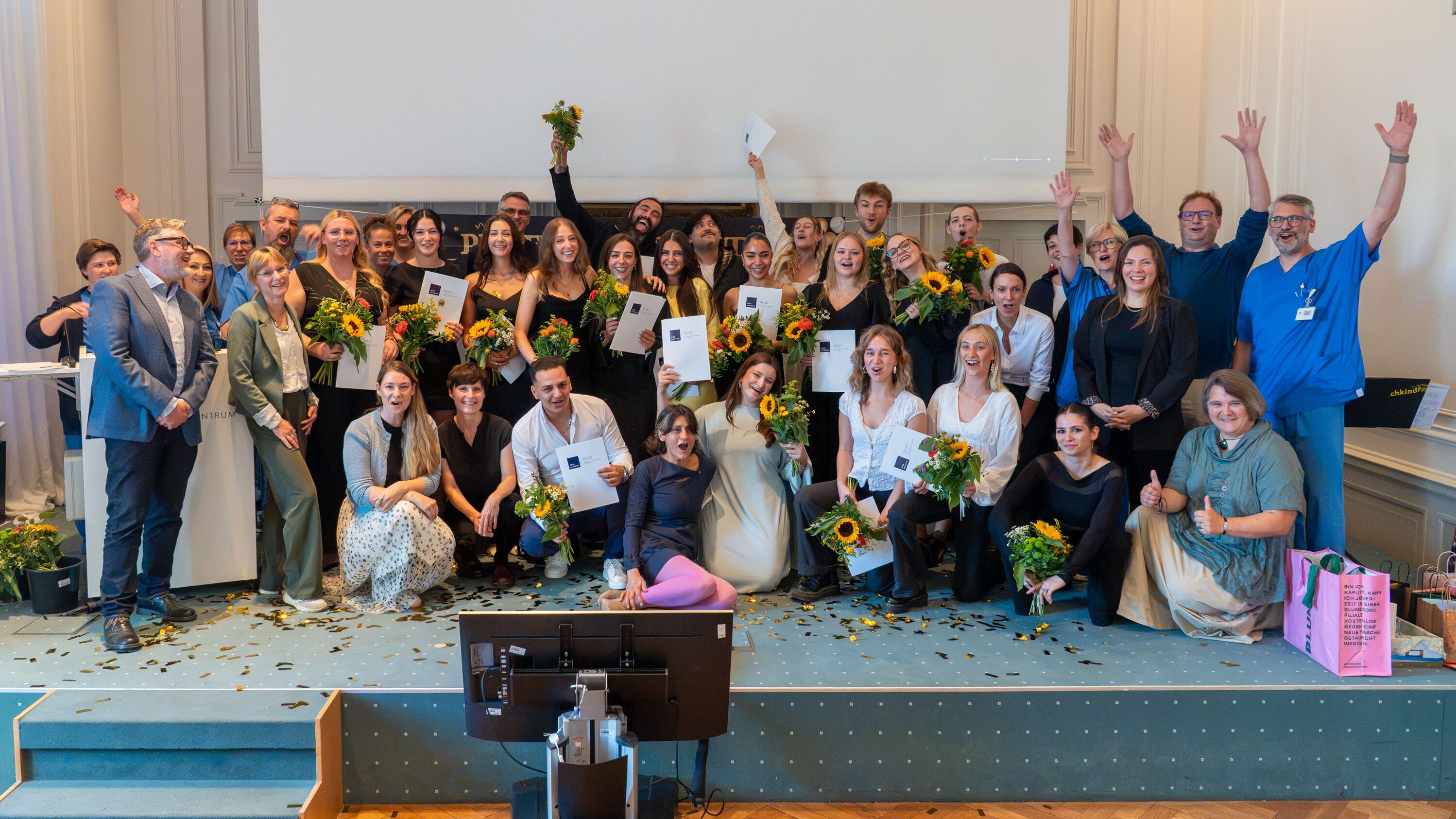 Eine Gruppe von etwa 40 Personen feiert zusammen auf einer Bühne. Viele halten Sonnenblumen und Urkunden in den Händen. Alle lächeln und zeigen Freude, einige winken in die Kamera. Der Hintergrund ist hell und die Atmosphäre wirkt festlich.