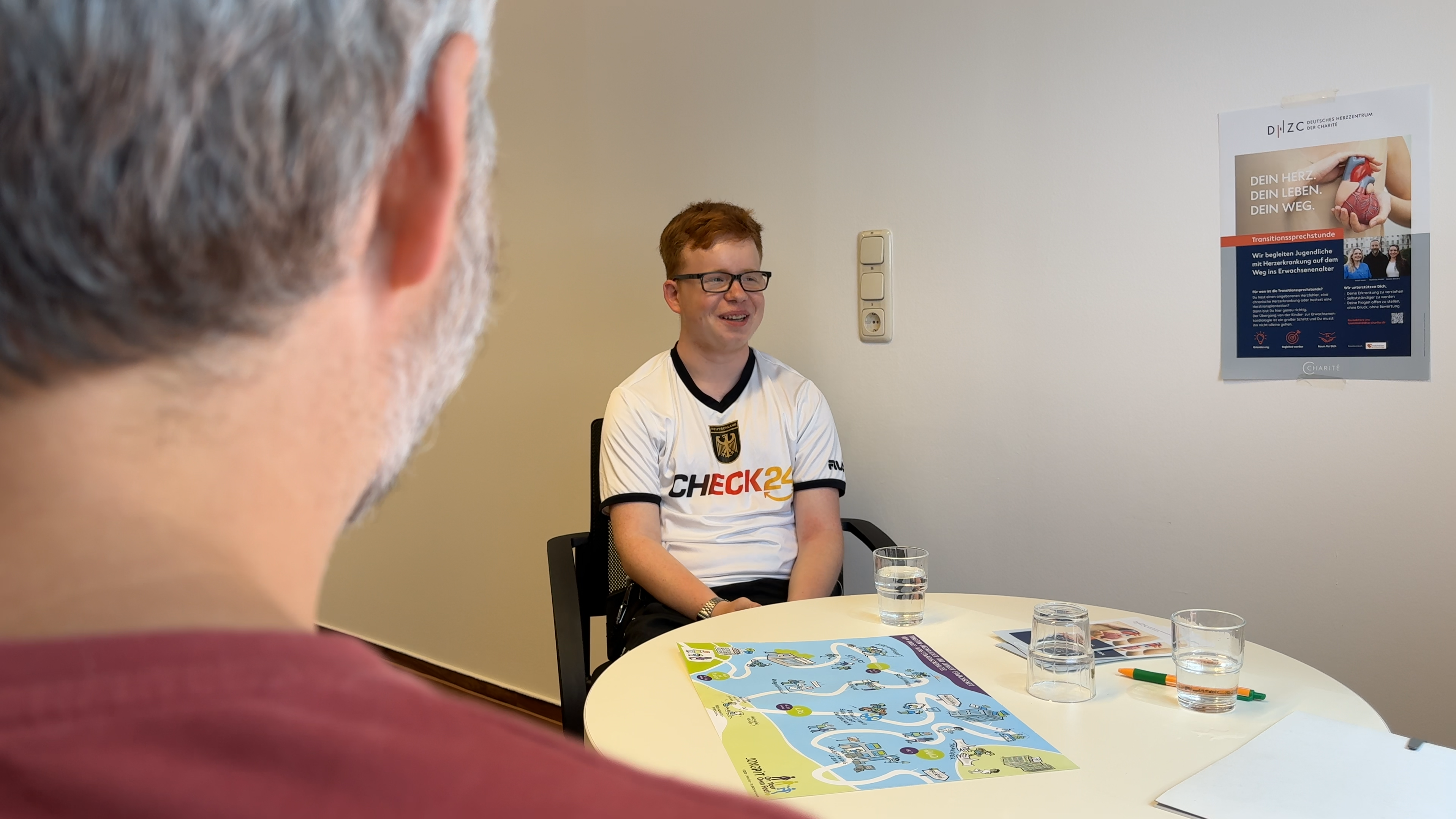Ein junger Mann mit Brille trägt ein T-Shirt mit der Aufschrift „CHECK24“. Er sitzt an einem Tisch, auf dem sich eine Karte und einige Gläser Wasser befinden. Sein Gesicht ist freundlich und er blickt in Richtung eines anderen Menschen, der von hinten zu sehen ist.