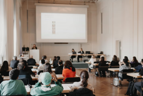 Ein Raum mit mehreren Personen, die in einem Vortrag sitzen. Vorne steht eine Rednerin an einem Podium, während eine Präsentation an die Wand projiziert wird. Die Zuhörer aus verschiedenen Altersgruppen und Berufen sind aufmerksam und notieren sich Informationen.