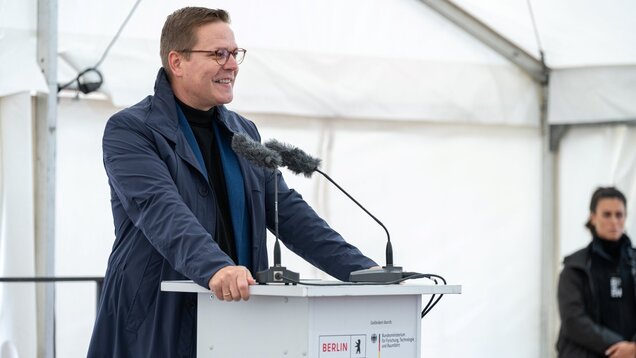 Ein Mann steht an einem Rednerpult, lächelt und spricht mit einem Mikrofon und einem Mikrofonarm. Im Hintergrund steht eine Person, die aufmerksam zuhört. Der Veranstaltungsort ist durch ein Zelt geschützt, und die Atmosphäre wirkt offiziell.