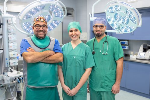Drei medizinische Fachkräfte in OP-Kleidung lächeln in einem Operationssaal. Sie tragen grüne Scrubs und passende Kopfbedeckungen. Im Hintergrund sind Operationslampen sichtbar. Die Atmosphäre ist professionell und freundlich.